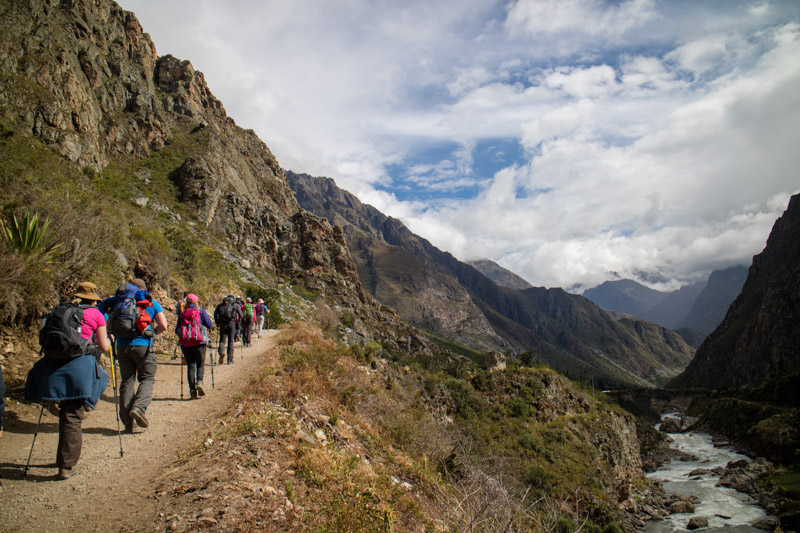 Hikers with backpacks walk along a narrow mountain path in a rugged landscape, surrounded by steep slopes and a river below, against a backdrop of partly cloudy skies.
